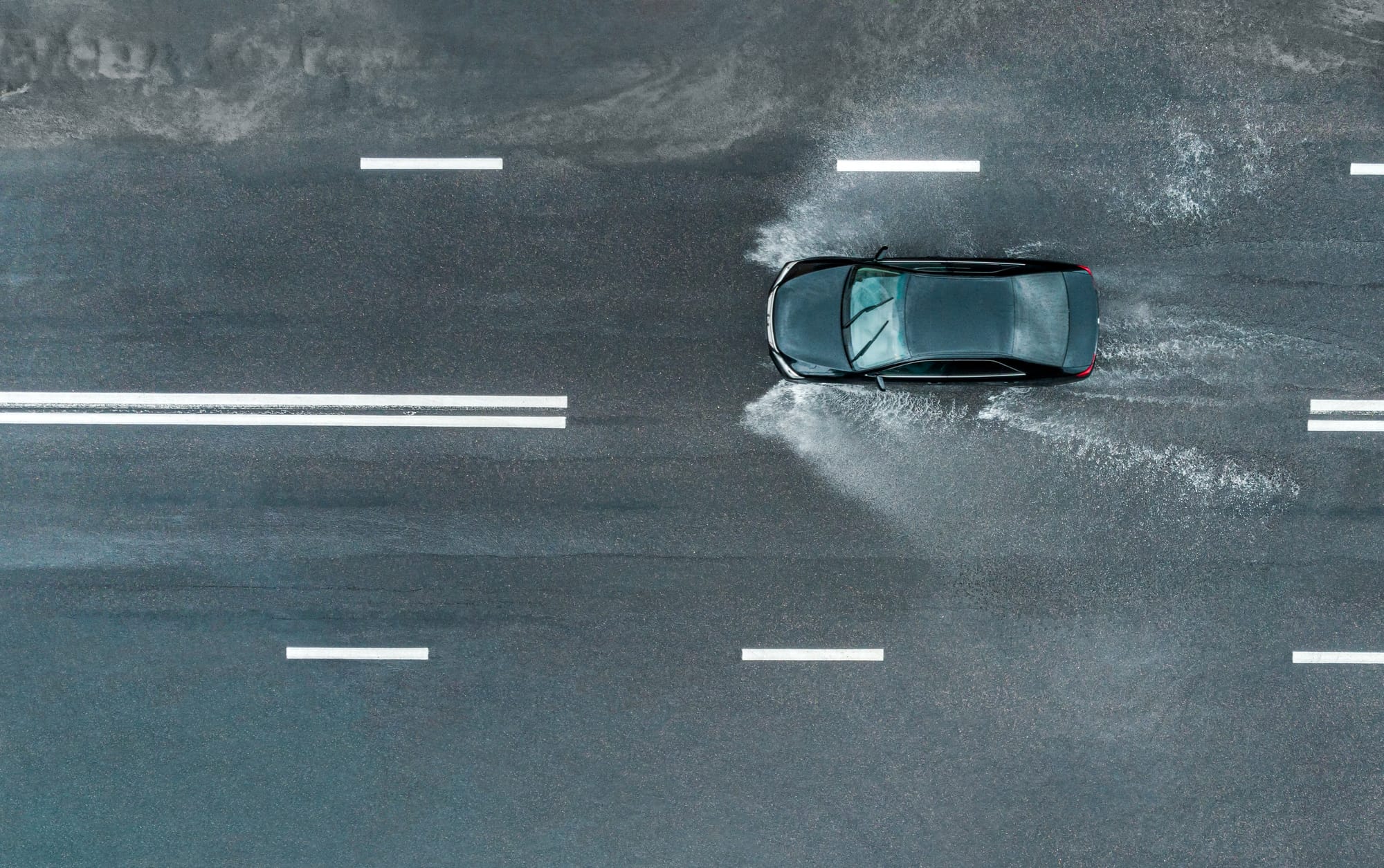 Car is driving fast with spray on a flooded road after a downpour. Aerial top view from flying drone.
