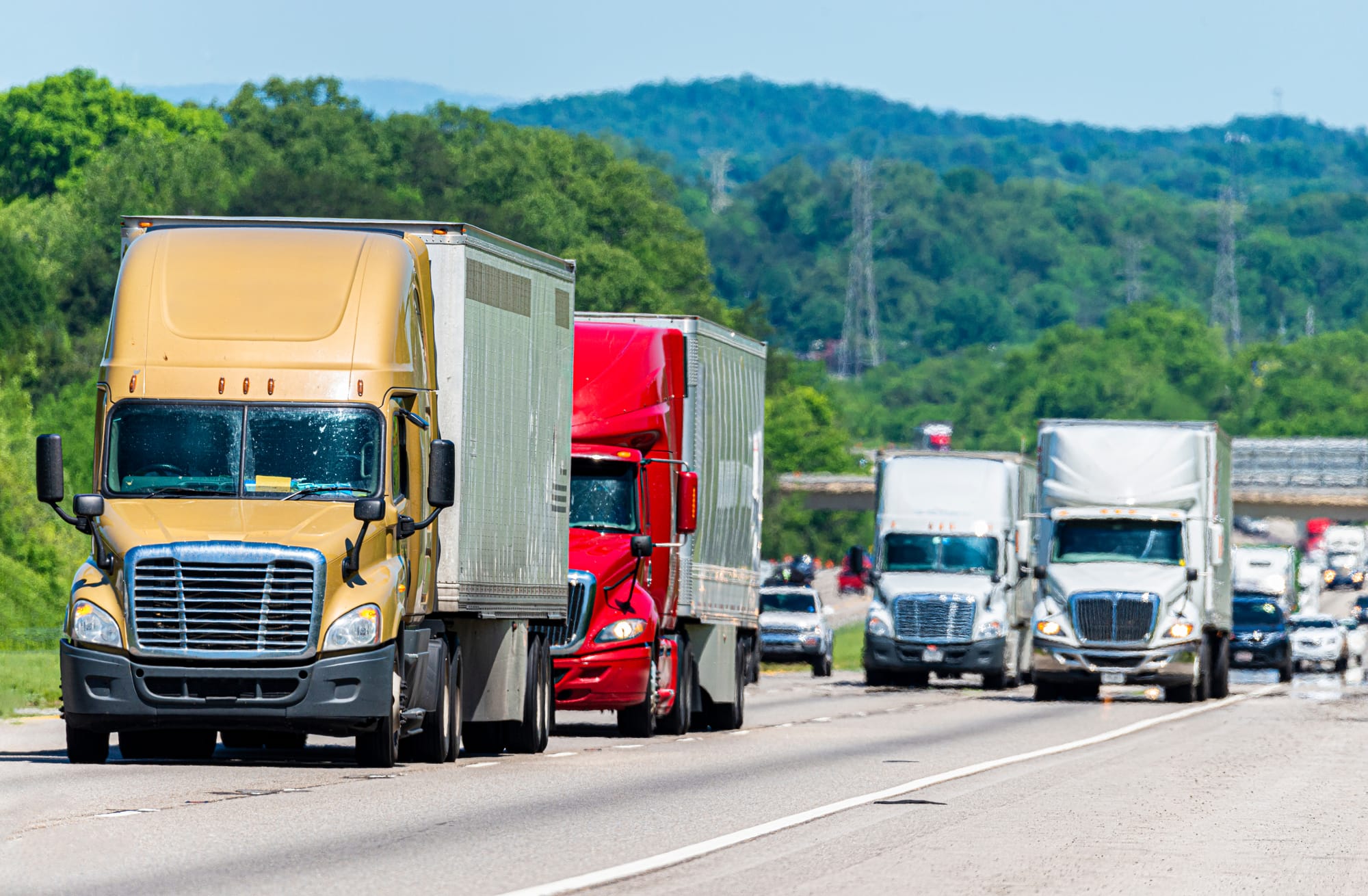 Lots of commerical trucks and other vehicles sharing a highway with green trees and hilly landscape in the background.